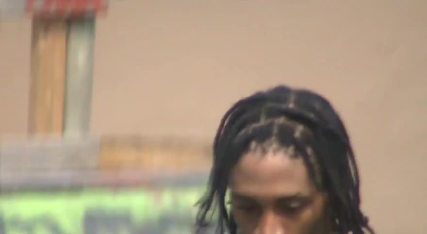 A raw, shaky, and blurry shot follows a man with dreadlocks as he walks past a wall