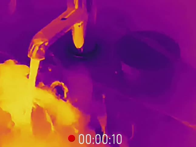 A thermal camera captures hot water running from a faucet as a person washes their hands in a sink, showing a vibrant display of heat signatures