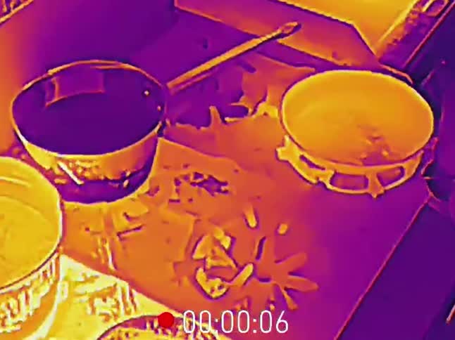 An infrared heat camera captures a kitchen stove with warm pots and pans in vibrant colors