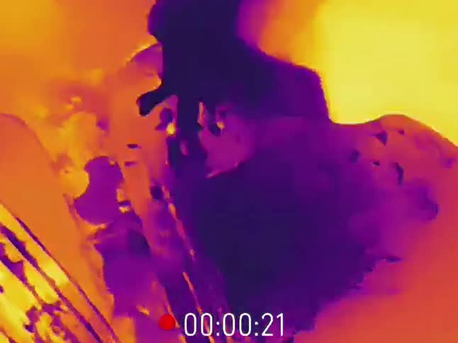 A first-person perspective from a thermal camera shows hands operating the controls of industrial machinery