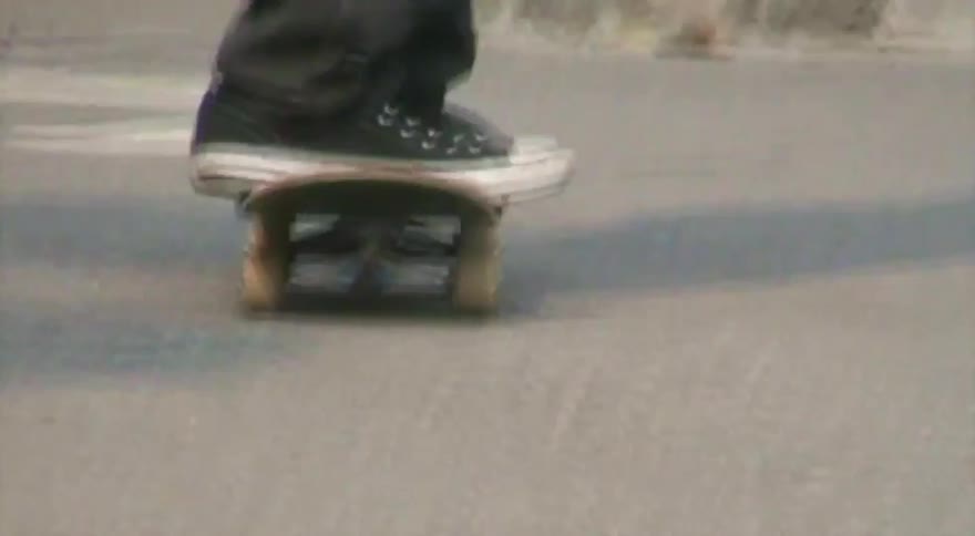 A stylized low-angle shot with a ghosting effect follows a person's feet as they ride a skateboard around a concrete skate park