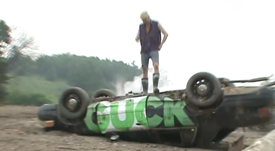 A low-angle shot zooms out from a man's muddy boots to reveal him standing on an overturned car in a rural landscape