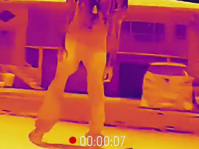 A skateboarder rides down the street at night and does a trick, captured on a thermal imaging camera