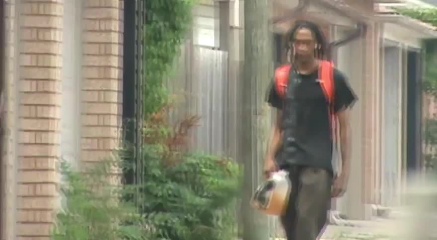 A young man with dreadlocks rides a skateboard down a sidewalk in a neighborhood while carrying a jug of juice