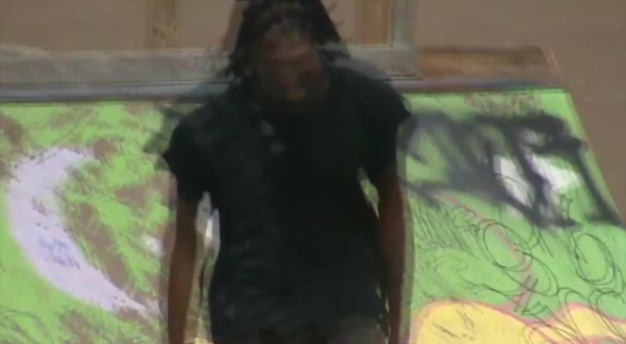A long-haired man skateboards through an urban park, cruising past graffiti-covered ramps before performing a jump