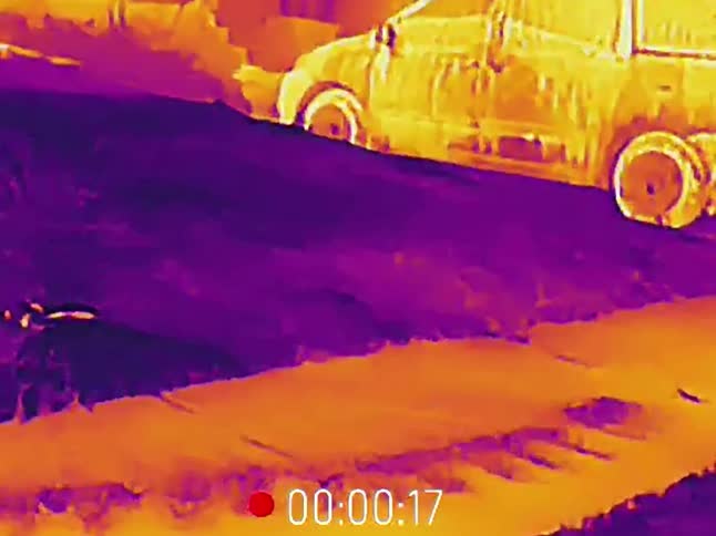 A man wearing a hat dances on the street at night, captured in a colorful thermal vision point of view