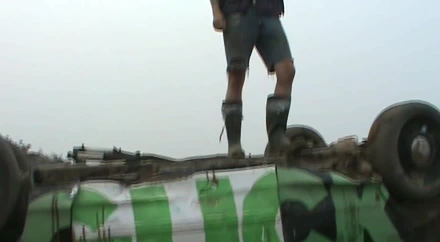 A man in sunglasses and rubber boots stands on top of a wrecked, overturned car with graffiti on its side