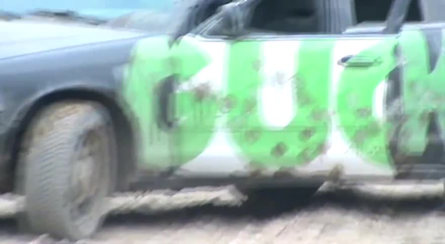 A cow grazes on a dirt patch next to a beat-up old sedan covered in green graffiti