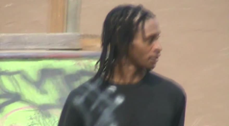 Vintage-style medium shot of a young Black man with braids carrying his skateboard and smiling while talking at a skatepark