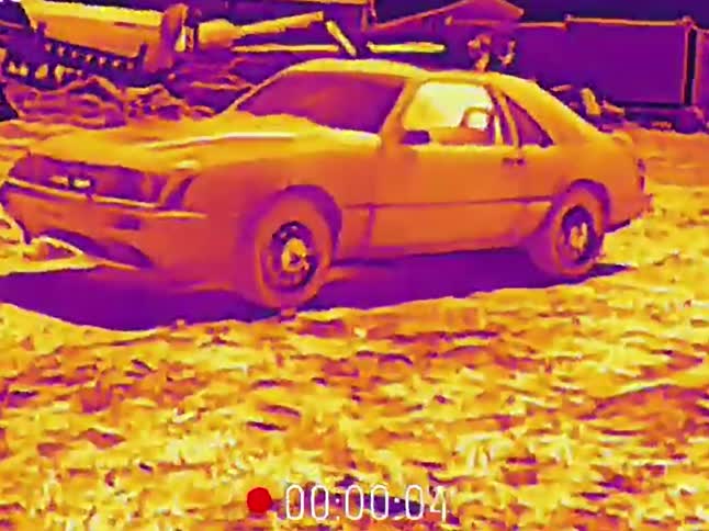 Surveillance-style thermal imaging footage of a vintage car abandoned in a junkyard