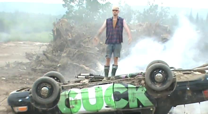 A slow zoom reveals a triumphant man with bleached hair and sunglasses standing on top of an overturned, smoking car in a junkyard