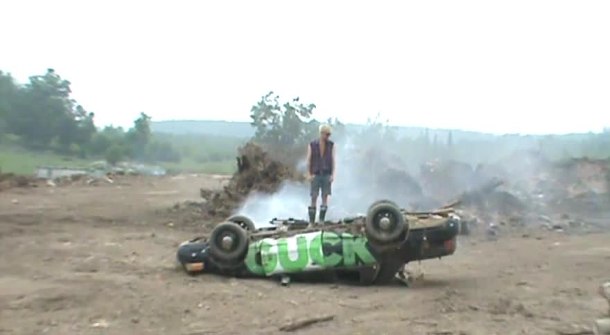A rebellious man with blonde hair and sunglasses stands victoriously atop an overturned, smoking car amidst a desolate junkyard