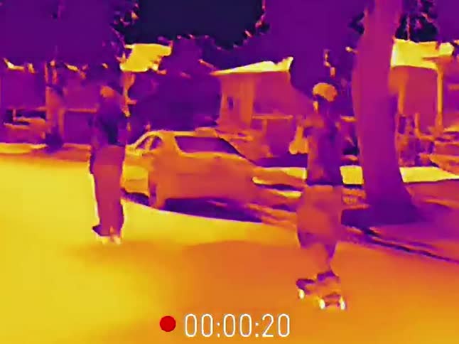 A thermal imaging view shows two friends skateboarding down a suburban street at night