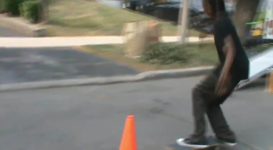 A skateboarder performs an ollie over an orange traffic cone and successfully lands the trick, knocking the cone over