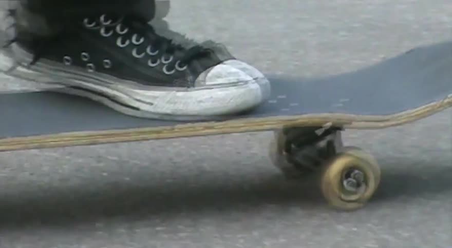 A low-angle close-up follows a skateboarder's feet as they ride and land a kickflip on a paved road
