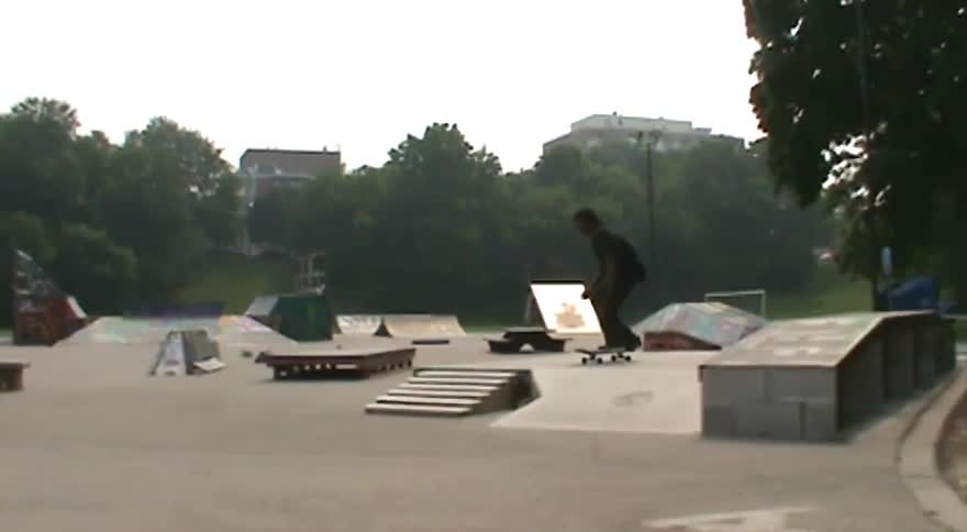 A skateboarder lands a trick at an outdoor skatepark in a wide-angle shot that zooms out to reveal the full scene