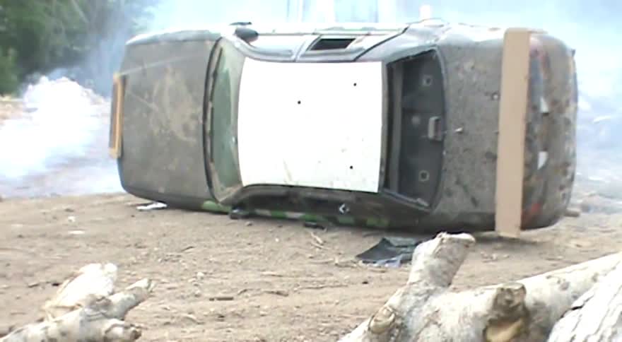 An overturned police car lies smoking on a dirt road after a violent crash