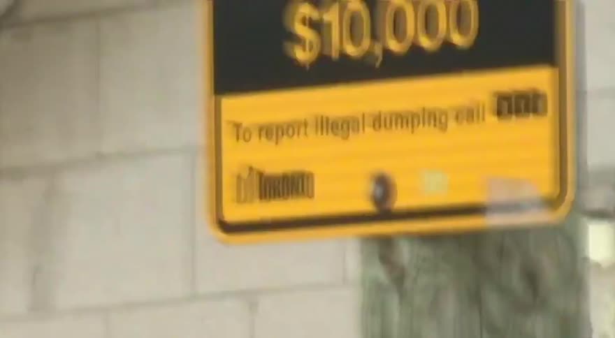 A yellow 'No Dumping' sign with a hefty fine warning hangs on a utility pole in a gritty urban back alley
