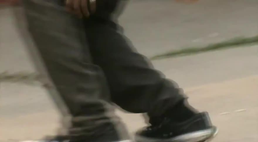 A skateboarder ollies over a fallen orange traffic cone in a gritty alley