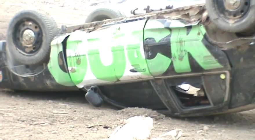 A close-up slow pan reveals an overturned and wrecked car with explicit green graffiti on its side at a junkyard