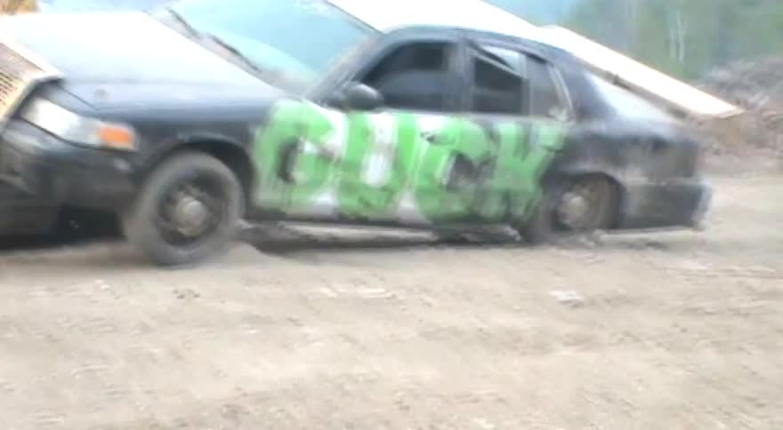 A bulldozer pushes a beat-up car with the word 'BUCK' spray-painted in green across a dirt yard