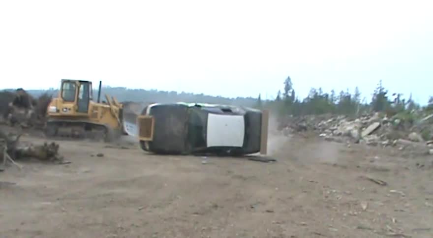 A bulldozer pushes a wrecked police car on its side across a dirt construction site