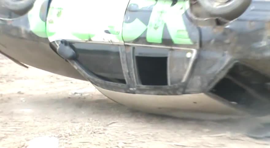 A stunt car with the word 'FUCK' spray-painted in green on its side flips over and lands on its roof in a junkyard
