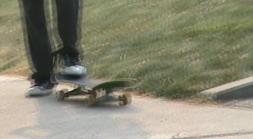 A young skateboarder in black sneakers flips his board on the sidewalk before the camera zooms in on his serious face
