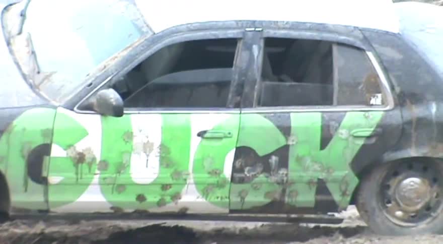 A heavy-duty bulldozer slowly crushes a dilapidated sedan covered in graffiti and bullet holes at a scrapyard