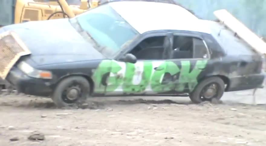 A modified demolition derby car with 'BUCK' spray-painted on the side is towed by a bulldozer through a muddy construction site