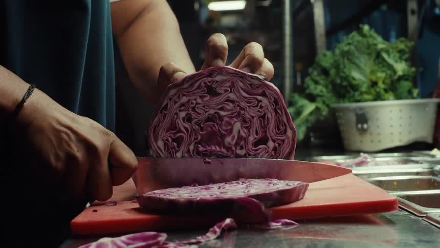 A chef's hands skillfully slice a fresh red cabbage on a red cutting board in a professional kitchen