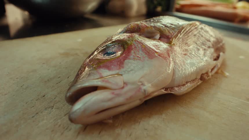 A sharp cleaver slices through a raw fish head on a wooden cutting board in a professional kitchen
