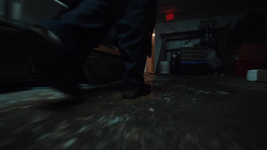 A creepy low-angle tracking shot follows a person walking through a dark and cluttered basement storage room