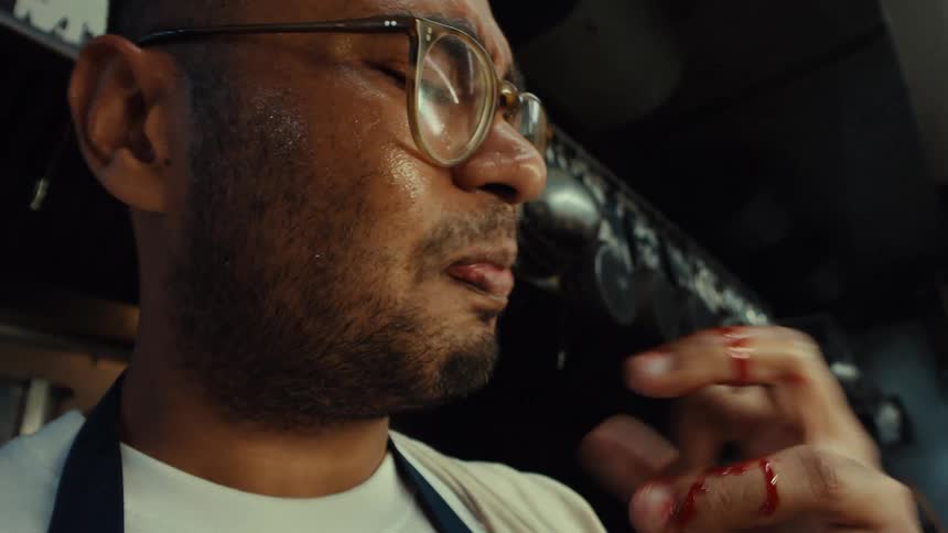 A sweaty chef in a hot kitchen accidentally cuts his finger and winces in pain while putting it in his mouth