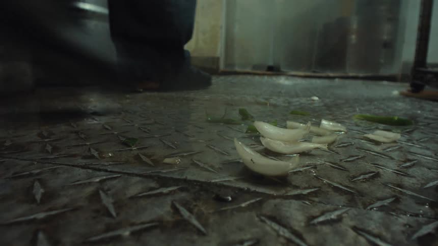 Sliced onions fall in slow motion onto the wet, metal diamond plate floor of a gritty commercial kitchen