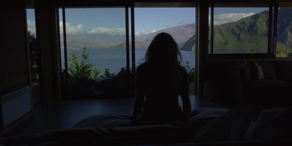 A woman sits down on a bed looking out a window