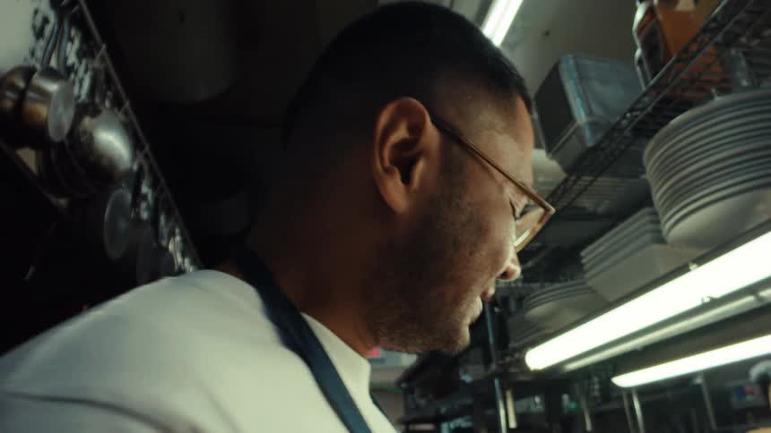A chef in a professional restaurant kitchen smiles as he tastes his culinary creation in slow motion