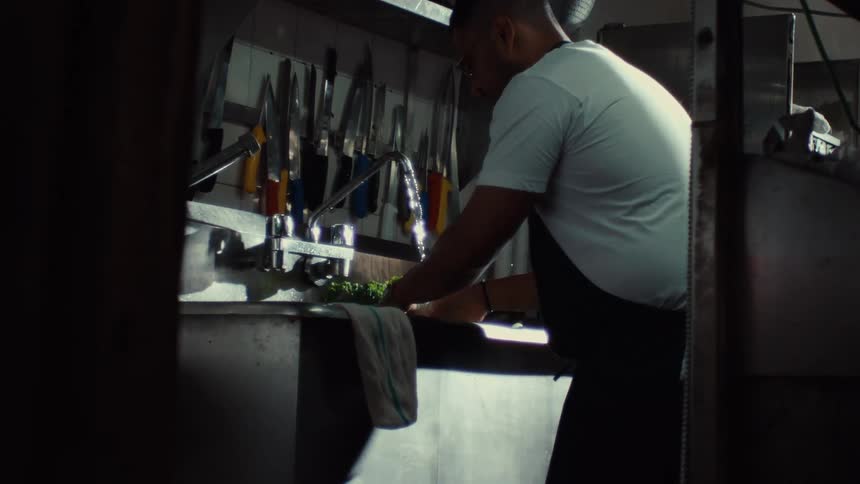 A cook in a professional kitchen carefully washes fresh green vegetables in a steel sink