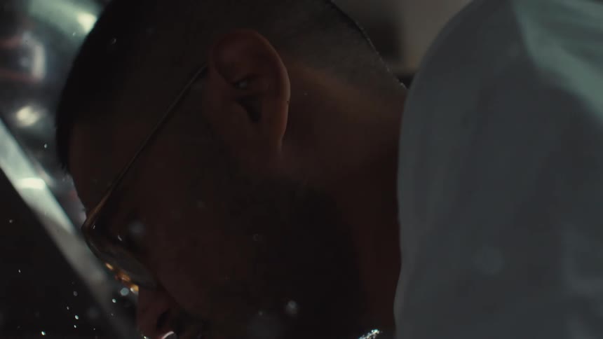 Dramatic slow motion close-up of a man's face as illuminated water droplets splash over him in a dark space
