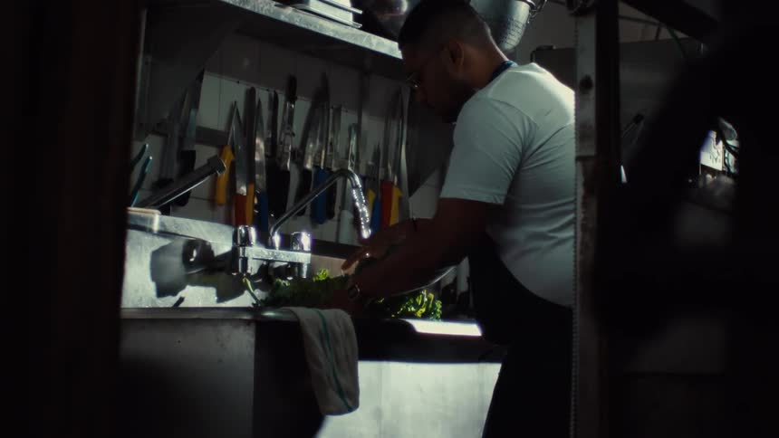 Slow motion view of a chef in a dimly lit professional kitchen meticulously washing fresh leafy greens in a sink