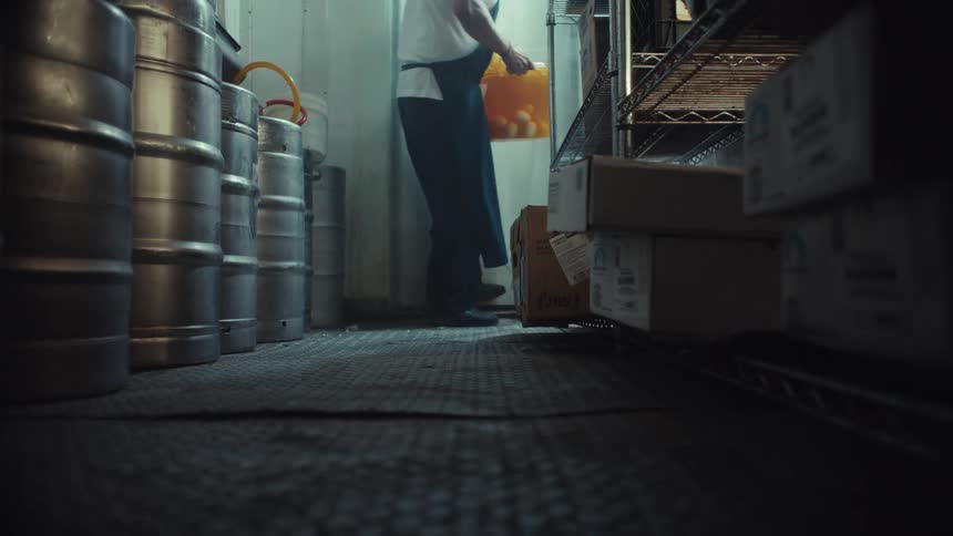 A low-angle view shows a kitchen worker walking through plastic curtains into a cold storage freezer to retrieve a bucket of ingredients