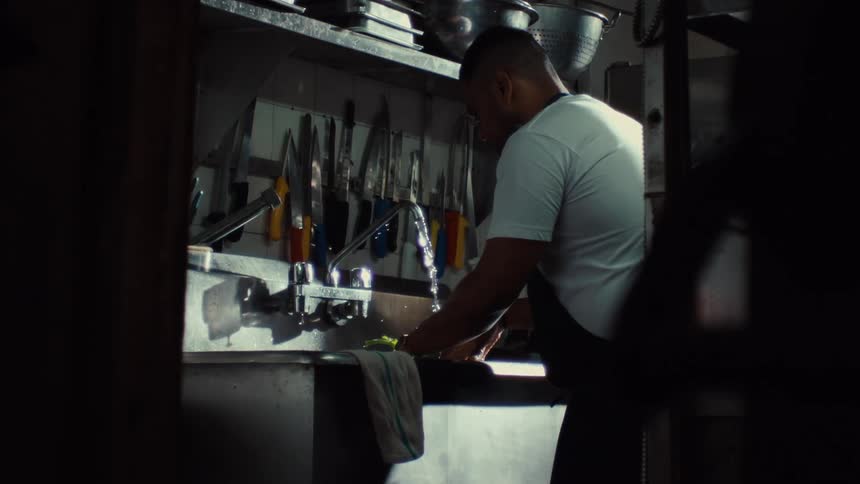 A chef works in a dimly lit professional kitchen, washing fresh leafy greens in a stainless steel sink in slow motion