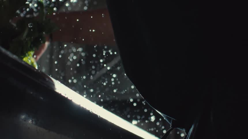 A cinematic slow-motion close-up of water droplets spraying off fresh greens being washed in a dark kitchen sink