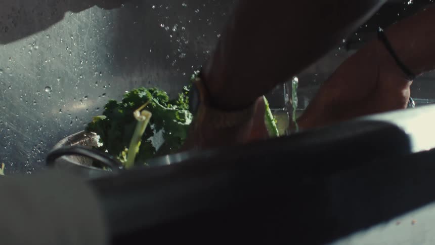 Fresh green kale is washed in a colander within a stainless steel sink, captured in dramatic slow motion