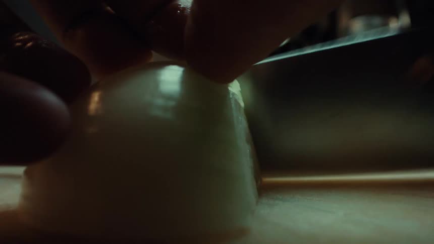 Extreme close-up macro shot of a chef's hands slicing a white onion on a cutting board in a dimly lit kitchen
