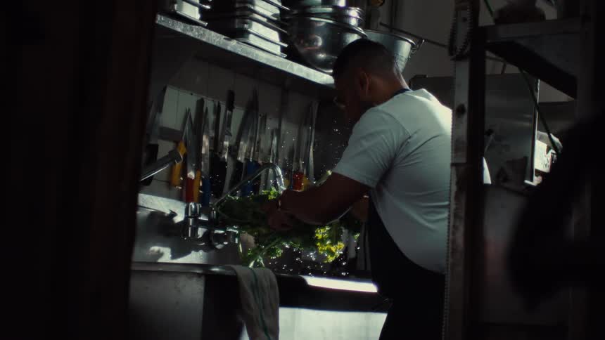 A professional chef methodically washes leafy greens in a restaurant kitchen sink in slow motion
