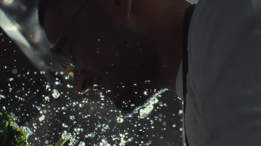 A man's face is backlit as he washes fresh produce, sending water droplets flying in cinematic slow motion