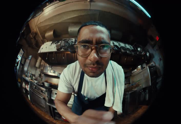 Fisheye lens POV of an intimidating chef in a professional kitchen who looks closely at the camera before swinging a large knife