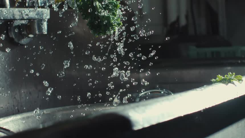 A close-up slow-motion shot shows water droplets splashing off of fresh green kale as it's washed in a stainless steel sink