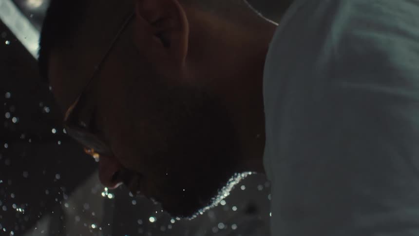 A dramatic slow-motion close-up from a low angle shows a man washing vegetables as water droplets spray and sparkle in the backlight
